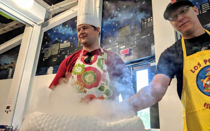 Two employees, both wearing aprons and one with a chef's hat, tend to a cauldron of dry ice.