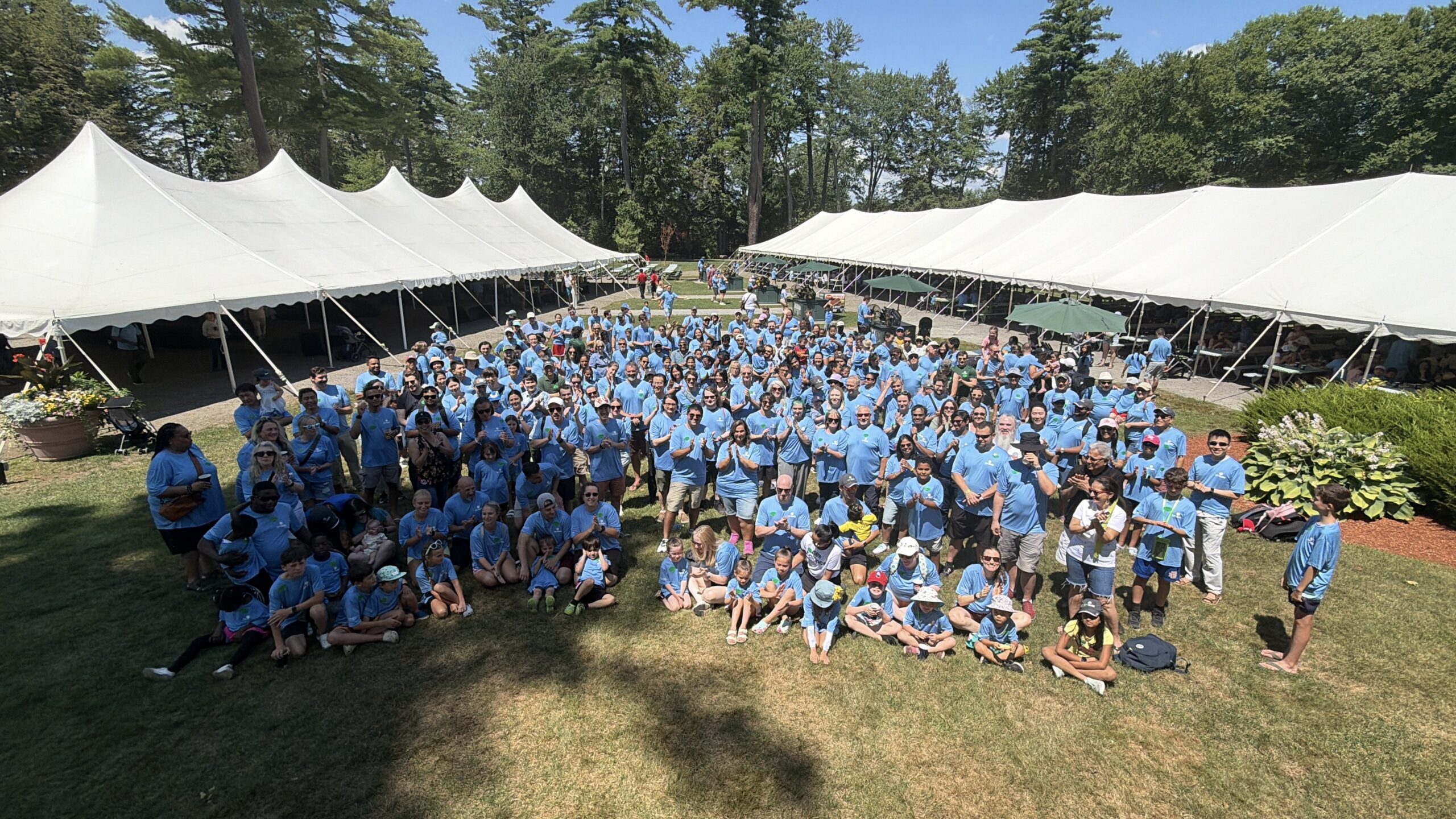 Large group of people in matching blue shirts posing together outdoors under white event tents in a park on a sunny day.