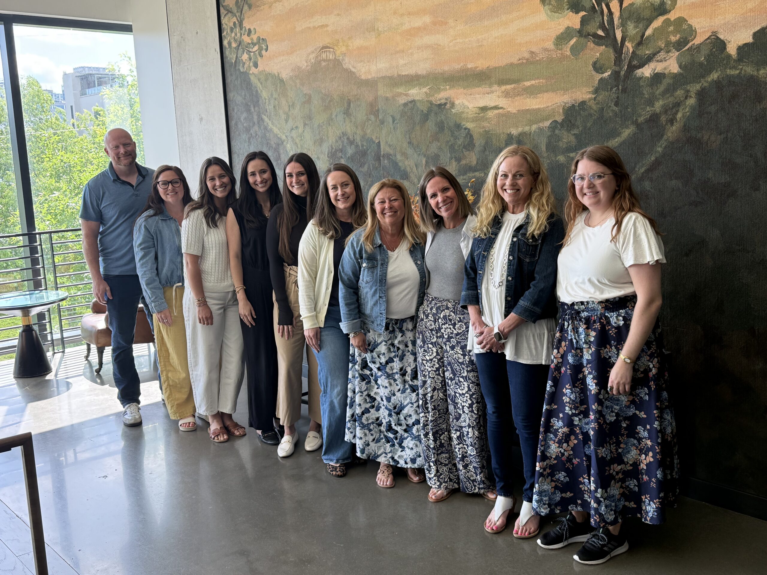 Group of eleven adults posing together indoors, smiling, in front of a large mural wall.