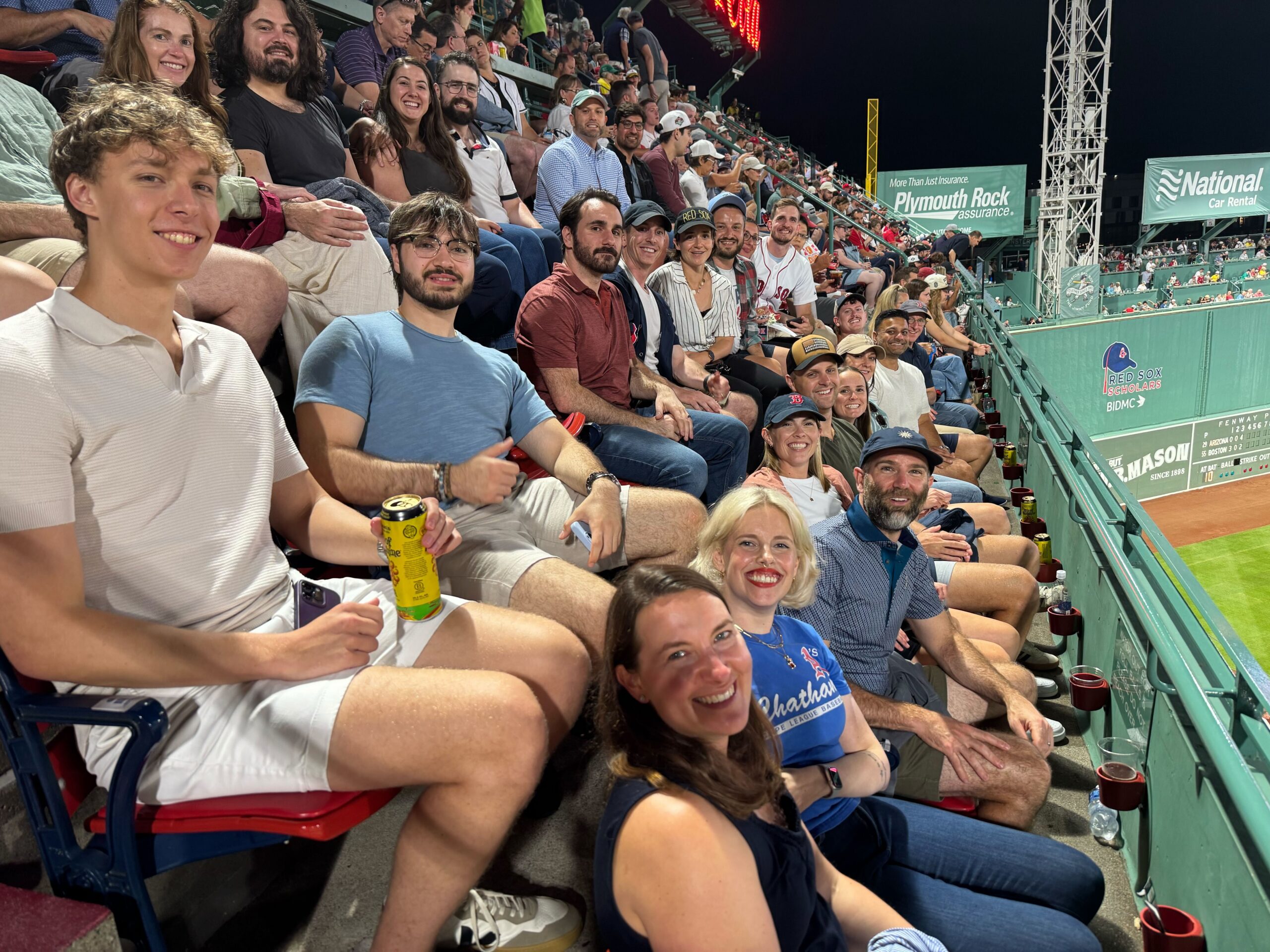 Smiling spectators seated in a baseball stadium at night, with banners and a field visible to the right.