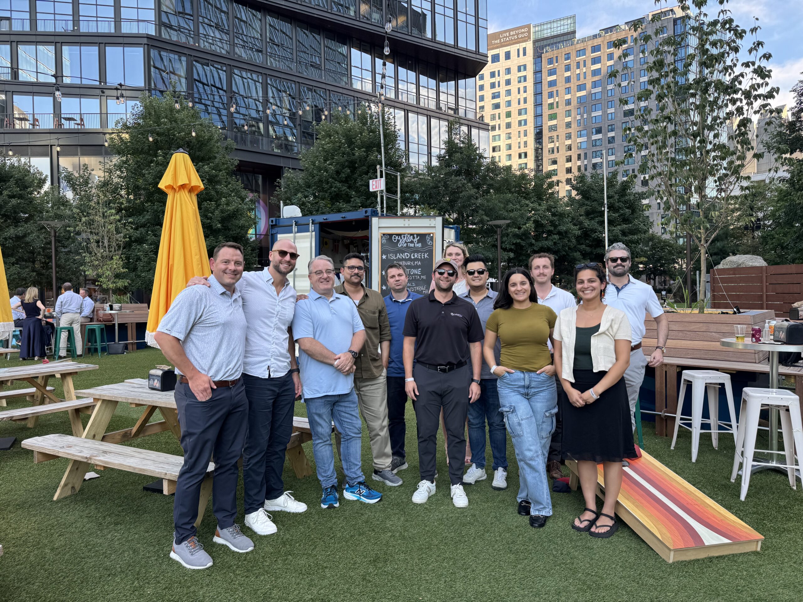 Group of coworkers posing for a photo in an outdoor urban courtyard with string lights and modern glass buildings in the background.