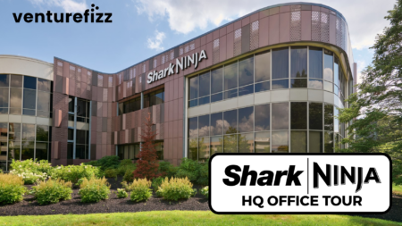 Exterior view of SharkNinja headquarters: curved glass facade, brown metal panels, and well-kept landscaping; Venturefizz logo in the top-left corner; foreground shows a large sign reading 'SharkNinja HQ OFFICE TOUR'