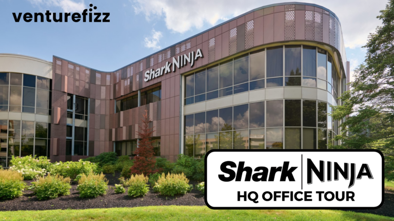 Exterior view of SharkNinja headquarters: curved glass facade, brown metal panels, and well-kept landscaping; Venturefizz logo in the top-left corner; foreground shows a large sign reading 'SharkNinja HQ OFFICE TOUR'