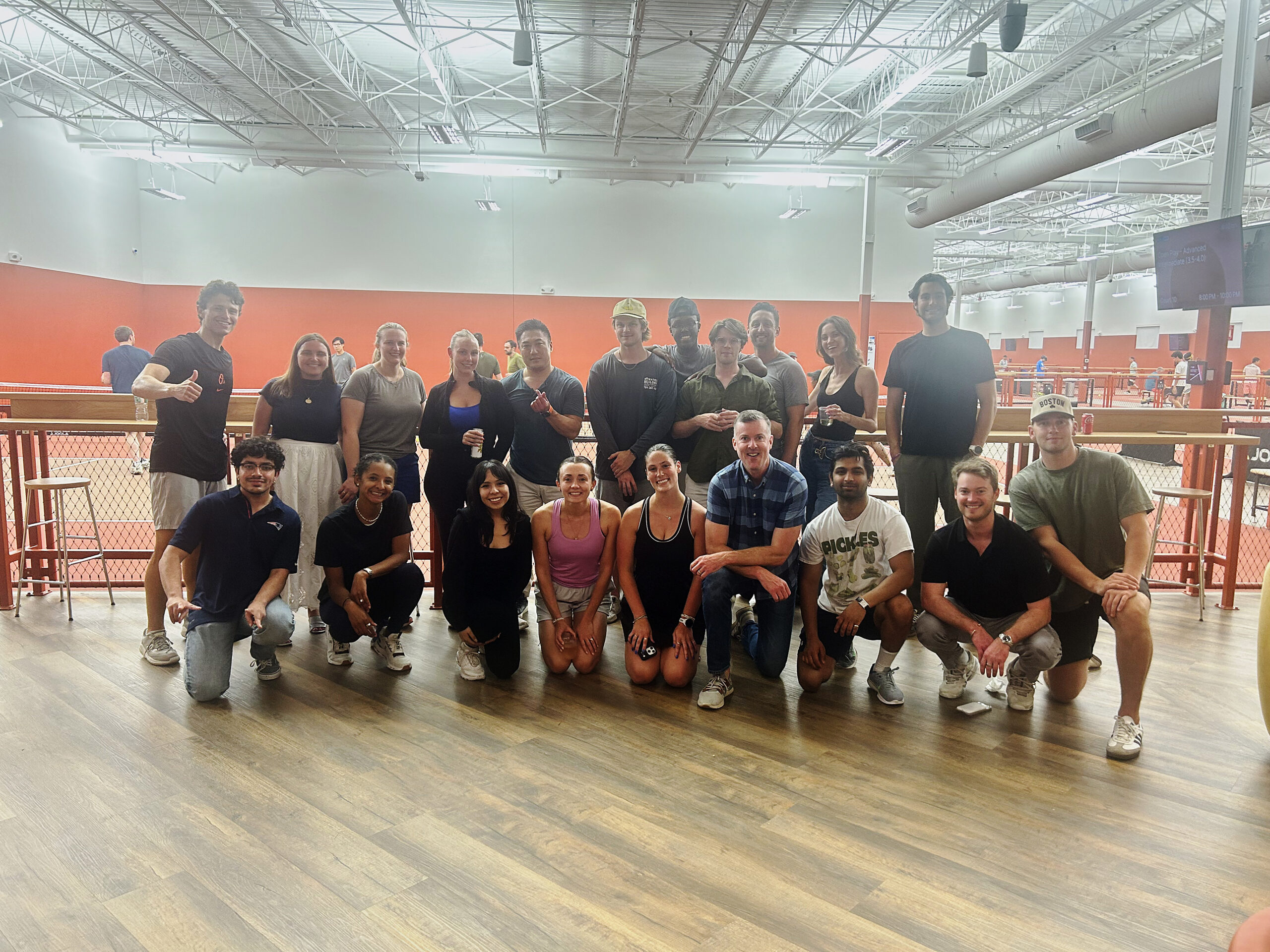 Group of smiling young adults posing for a photo inside an indoor gym, with an orange wall and tennis courts in the background.