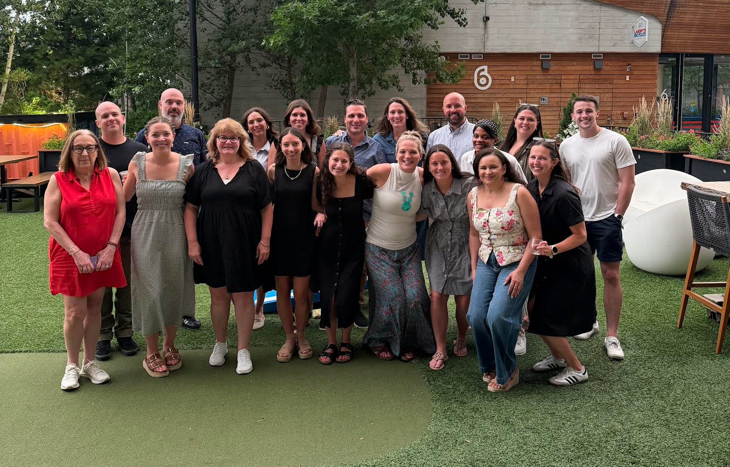 Group of about 20 adults posing together outdoors on a green artificial lawn, smiling for a group photo with a building and planters in the background.