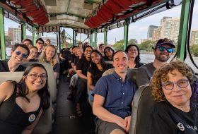 Group of smiling adults on a city sightseeing boat with skyline in the background.