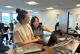Two women collaborate at a desk, one points at a laptop while a large monitor shows data in a bright office.