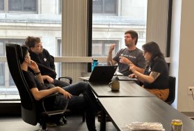 Four people sit around a long conference table in a bright office, engaged in conversation; one gestures with a hand while others listen with laptops nearby.