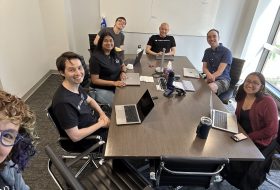 Six colleagues sit around a long conference table in a bright office, smiling at the camera; laptops, a microphone, and notepads lay on the table as a whiteboard with a chart is visible in the background.