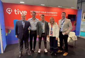 Five professionals posing at a trade show booth with a red backdrop and company branding behind them, smiles for the camera.
