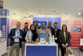 Group of eight professionals posing at a trade show booth for the company 'tive', with banners, brochures, and branded water bottles on a blue front desk.
