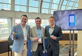 Three men in suits pose in a bright conference space, each wearing a badge and holding small blue devices.