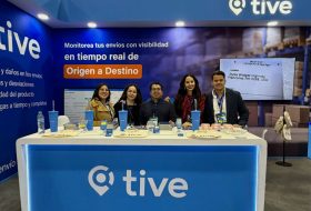 Five people stand behind a blue trade‑show booth with the 'tive' logo; banner reads 'Origen a Destino' and 'Monitorea tus envíos'.