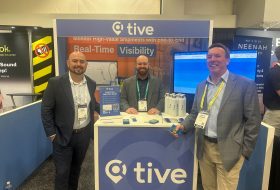 Three men in suits at a 'tive' trade show booth, displaying banners for Real-Time Visibility with branding in blue.