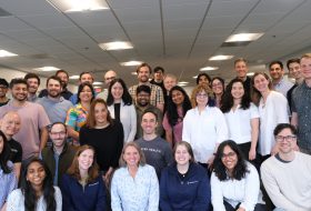 Large group of diverse coworkers posing together in an office corridor, smiling at the camera.