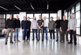 Twelve men standing in a row in a modern office lobby with large windows.