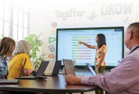 Woman presenter points to colorful bar charts on a large display as colleagues watch in a bright meeting room with laptops open on a round table.