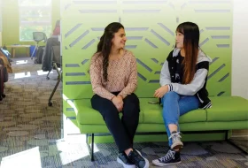 Two young women sit on a lime-green bench, facing each other and smiling while chatting in a modern lounge with glass partitions.