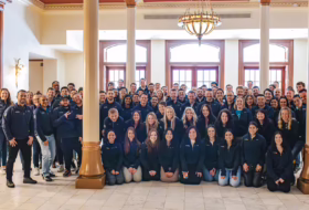 Large group of employees in navy jackets posing for a team photo in a bright lobby with columns and a chandelier, indicating a company gathering.