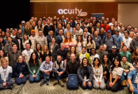 A large group of diverse people posing for a company group photo in a conference room, with the 'acuityMD' logo on the back wall.