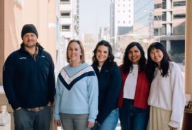 Group of five adults standing on a city street, smiling at the camera with urban buildings in the background.