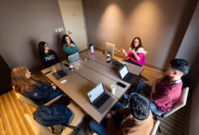 Seven colleagues sit around a rectangular conference table in a beige-walled room, with laptops, cups, and bottles; a woman in a pink sweater gestures as she speaks.