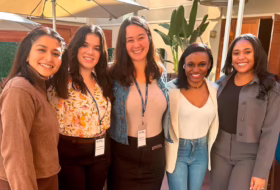Five young women standing together outdoors at an event, wearing name badges and smiling at the camera.