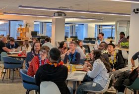 Group of coworkers at a casual office party, wearing colorful leis, seated around tables with drinks and snacks.