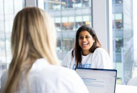 Smiling woman with dark hair talks with a colleague in a bright office; the back of another person’s blond hair and a laptop are visible in the foreground.