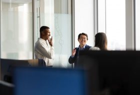 Three coworkers chatting near a glass wall in a bright office, smiling and engaged in conversation.