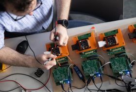 Person wearing a plaid shirt tests circuit boards with a handheld probe, wiring and USB cables visible on a workbench.