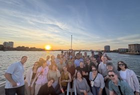 A large group of people on a boat posing for a selfie at sunset with a city skyline in the background.