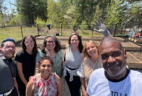 Smiling group of adults taking a selfie in a backyard garden with raised wooden beds and a fence in the background, a gloved hand raised on the right.