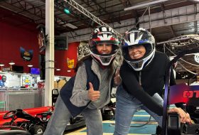 Two people wearing full-face helmets lean toward the camera and give thumbs up in an indoor go-kart facility.
