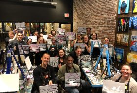 Group of smiling people in a brick-walled art studio posing with their painted canvases showing trees and bridges during a painting class.