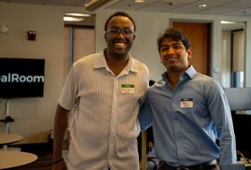 Two smiling men with name tags stand together in a corporate office lobby.