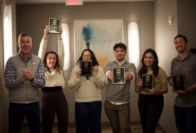 Six adults in a hallway posing with award plaques and smiling for the camera, celebrating achievements in an office setting.
