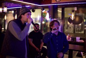 Two male performers sing into wireless microphones on a dimly lit bar stage, with colorful neon lights in the background.