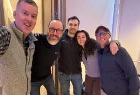 Five adults posing for a group selfie in a hallway, smiling at the camera.