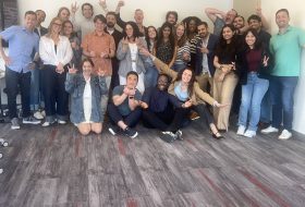 Group of coworkers posing together in an office, smiling and making playful hand gestures.