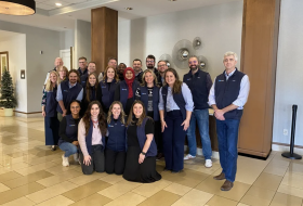 Group of about 20 coworkers in matching navy vests posing for a photo in a hotel lobby with tiled floor and light-colored walls.