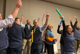 Group of people raising colorful foam tubes in a celebration indoors, smiling and cheering.