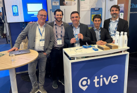 Five men in business attire pose at a trade show booth with a blue 'tive' counter; one man holds a device, others smile behind the table of brochures and water bottles.