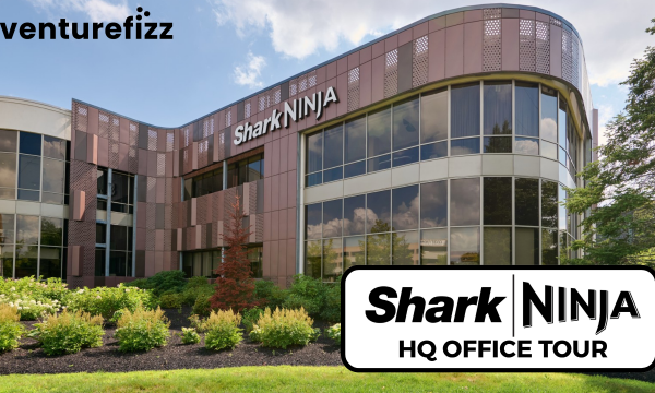 Exterior view of SharkNinja headquarters: curved glass facade, brown metal panels, and well-kept landscaping; Venturefizz logo in the top-left corner; foreground shows a large sign reading 'SharkNinja HQ OFFICE TOUR'