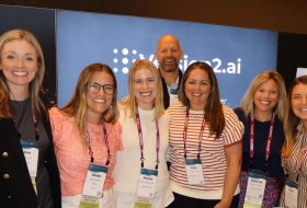 Group of seven smiling professionals wearing conference lanyards posing for a photo in front of a blue event backdrop with branding.