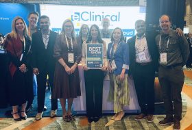 Group of ten professionals at a conference posing together, with a central sign reading 'BEST OF SHOW' held by two women, in front of a bright display.