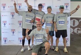 Five runners in gray t-shirts with 'eClinical' logo pose in front of a Home Base backdrop, wearing race bibs.